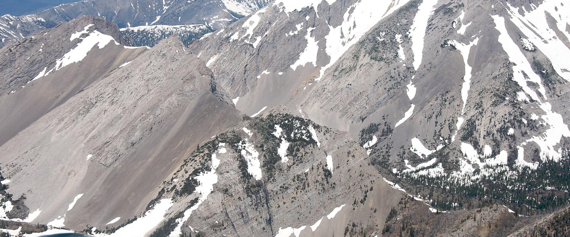 A high-altitude aerial view of rugged, snow-dusted mountain peaks with a small alpine lake nestled in a valley below.