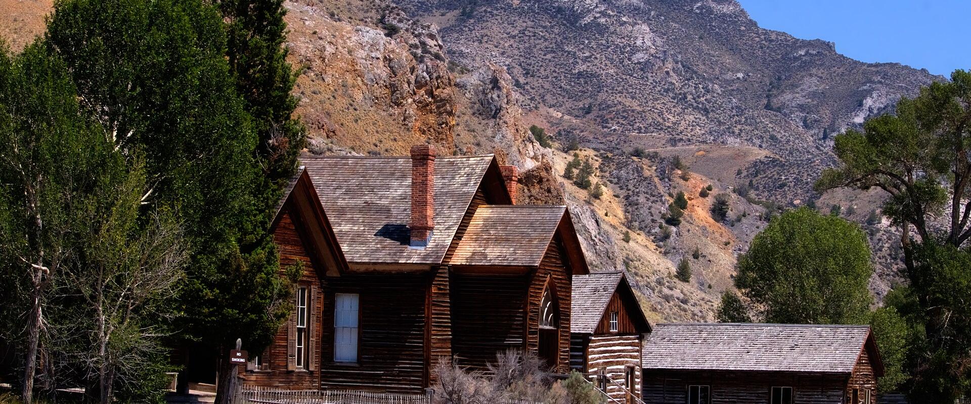 The image shows a historic log cabin with a steep roof and chimney, surrounded by a wooden fence and trees, set against a backdrop of rugged, arid mountains under a clear blue sky.