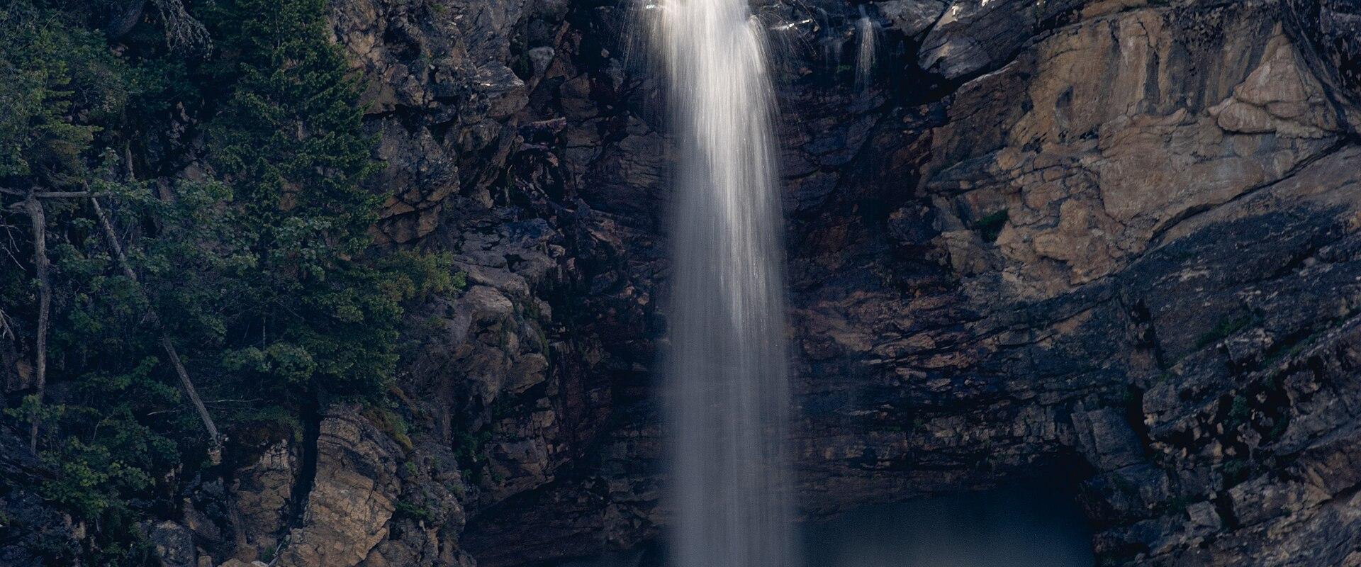 A long-exposure photograph of a waterfall cascading down a rocky cliff face into a pool, surrounded by dense forest and trees.