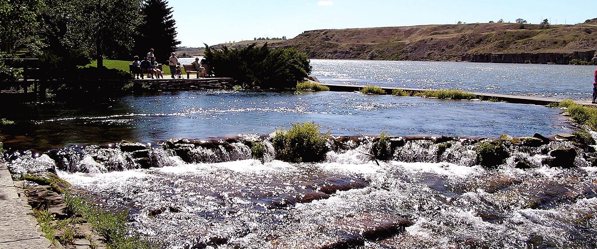 The image shows a scenic view of a river with a small weir creating white water, a calm lake in the background, and people sitting on a bench near the water's edge under trees.