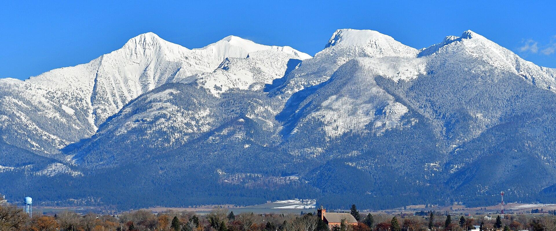 The image shows a wide landscape view of a small town with a prominent church steeple in the foreground, set against a backdrop of snow-capped mountains under a clear blue sky.