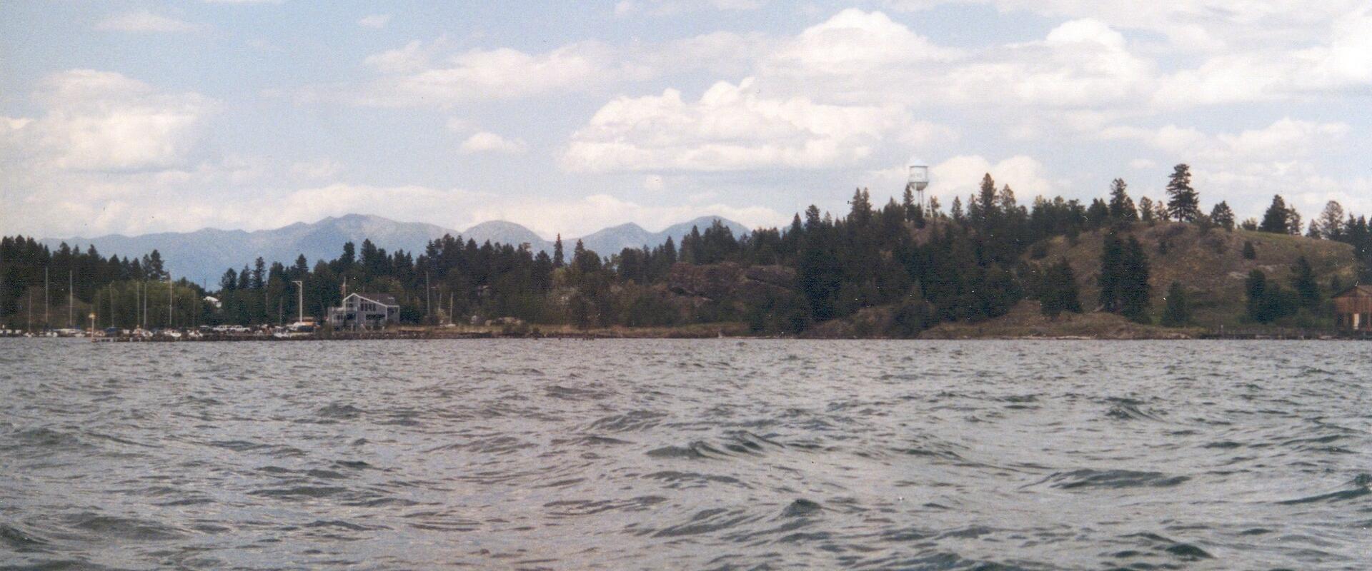 The image shows a large body of water with a forested shoreline, distant mountains, and a small structure on the shore under a partly cloudy sky.