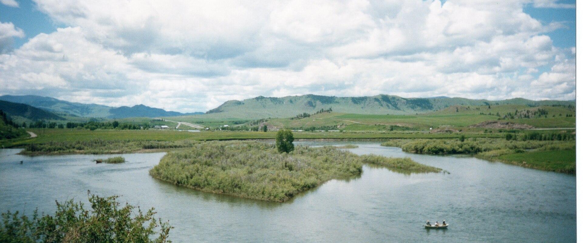 The image shows a wide river with a grassy island in the center, rolling green hills in the background, and a small boat on the water under a cloudy sky.