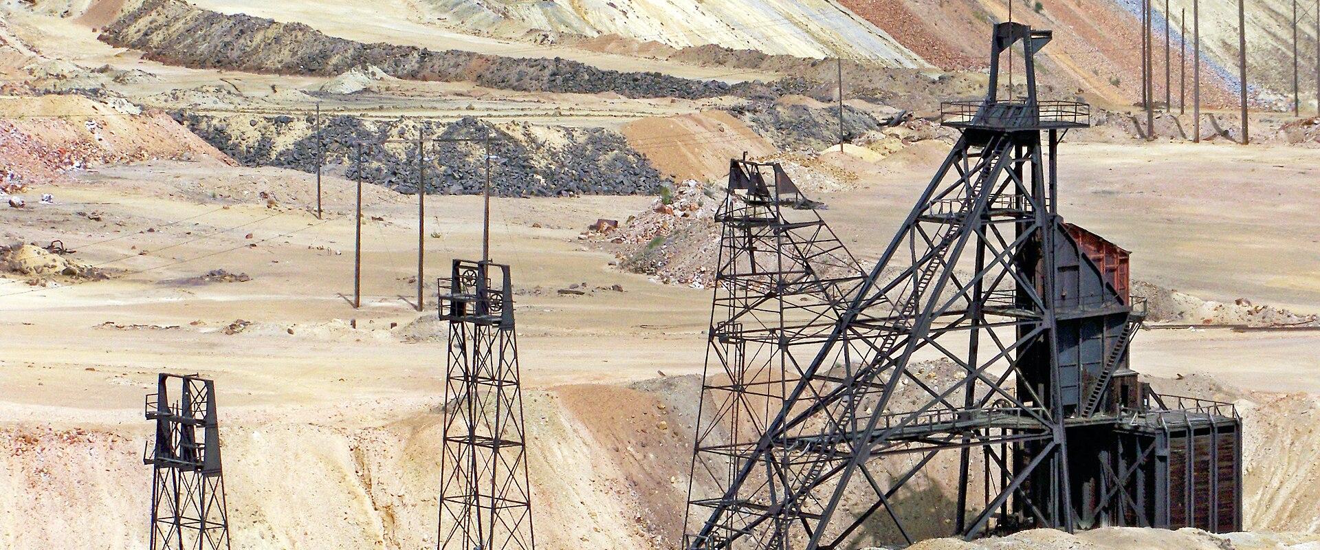 The image shows a large, rusted steel headframe and associated mine structures set against a backdrop of a massive, terraced open-pit mine with colorful mineral deposits.