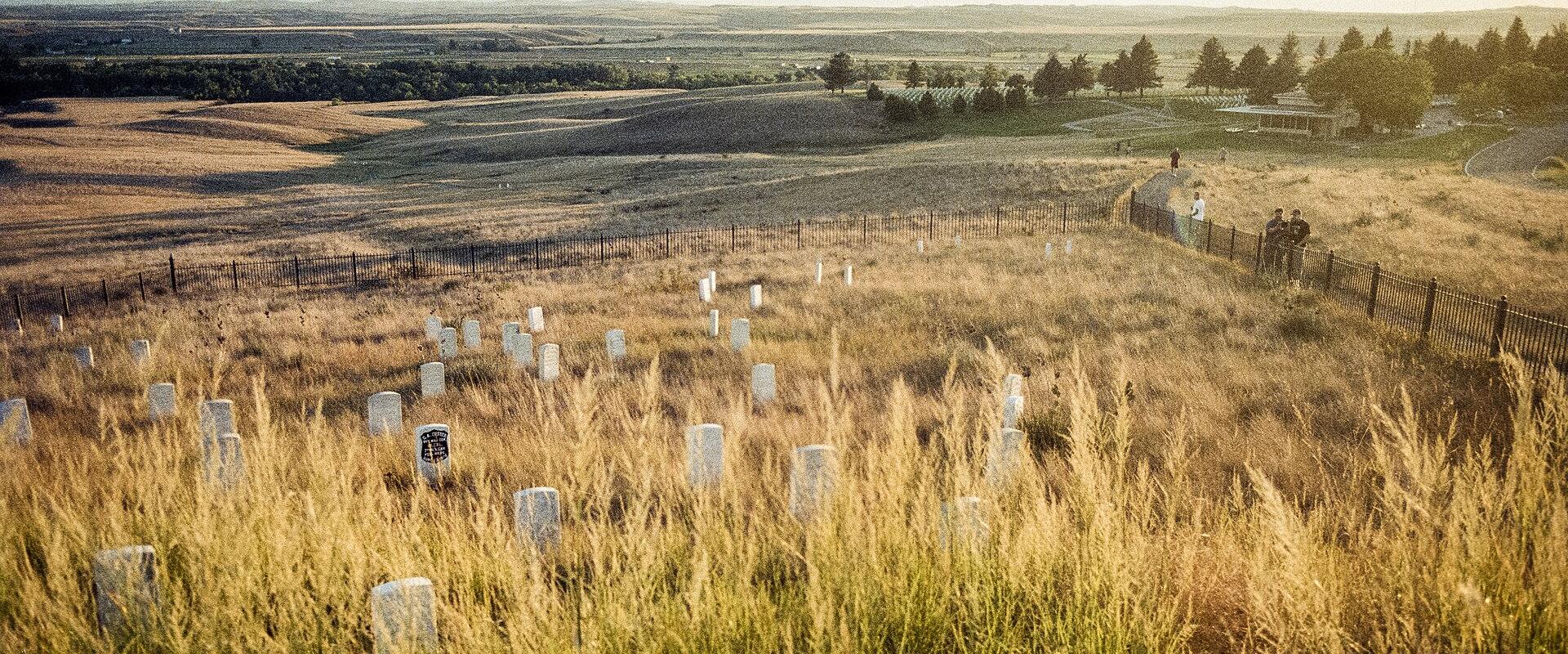 The image shows a wide, grassy battlefield landscape with numerous white headstones scattered across the foreground, a fence line, and a few visitors in the distance under a bright sky.